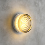 Clear rippled-glass Clérachic wall light with a pale tint at the rim, glowing warm on a grey concrete wall.