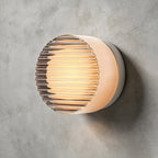 Round Clérachic wall light with a ribbed clear glass face and white sides, glowing warm on a concrete wall.
