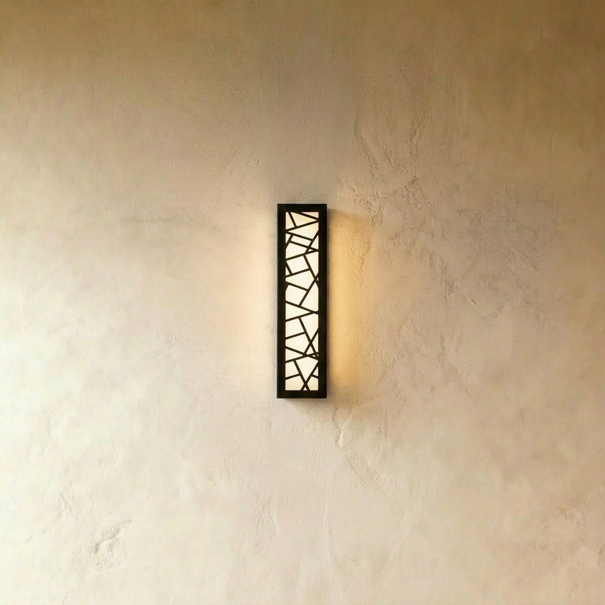 Clérachic tall narrow black wall lamp with a cut-out geometric pattern, glowing warm white against a pale limewash wall.