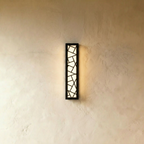 Clérachic long black wall lamp with a geometric cut-out front, lit warm white and casting a soft glow on a textured wall.