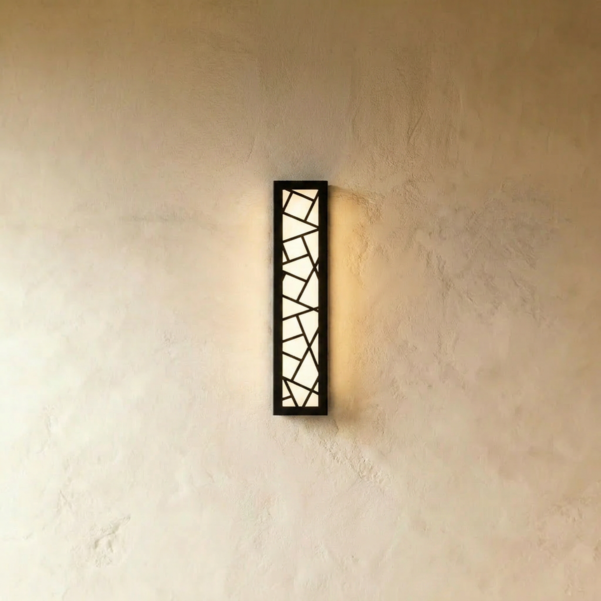Clérachic long black wall lamp with a geometric cut-out front, lit warm white and casting a soft glow on a textured wall.