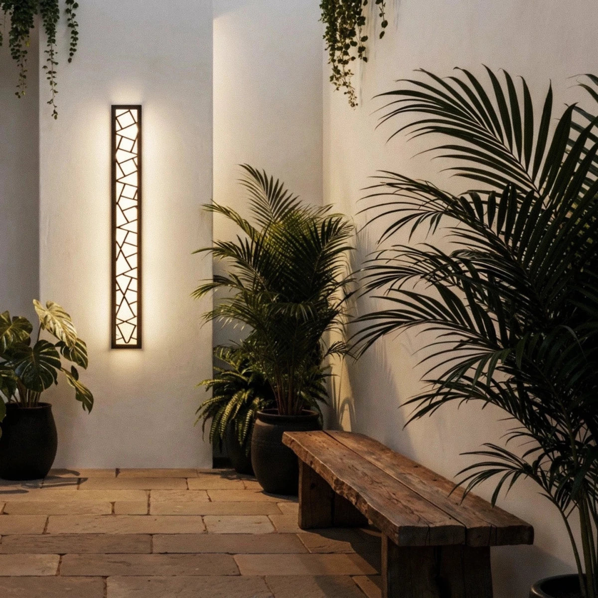Clérachic tall black cut-out wall lamp glowing beside a stone-tiled courtyard, with a rustic wooden bench and potted palms.