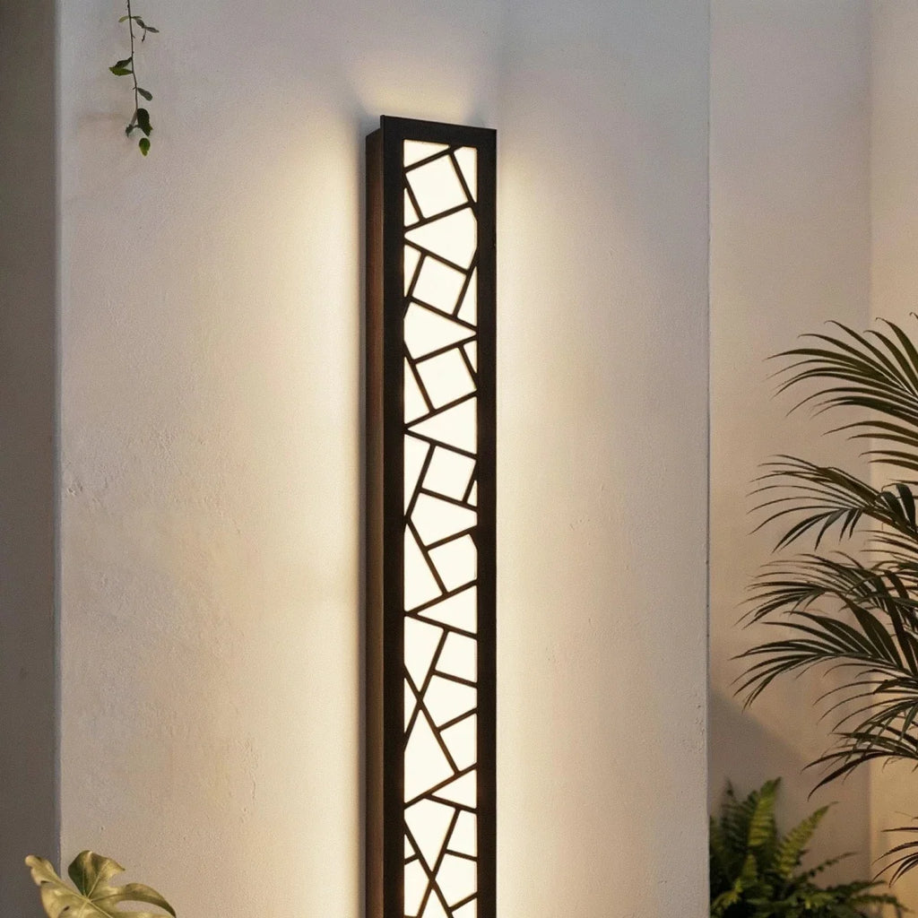 Clérachic tall black wall lamp with a geometric cut-out grille glowing warm white on a white wall among palm leaves.
