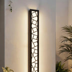 Clérachic tall black wall lamp with a geometric cut-out grille glowing warm white on a white wall among palm leaves.