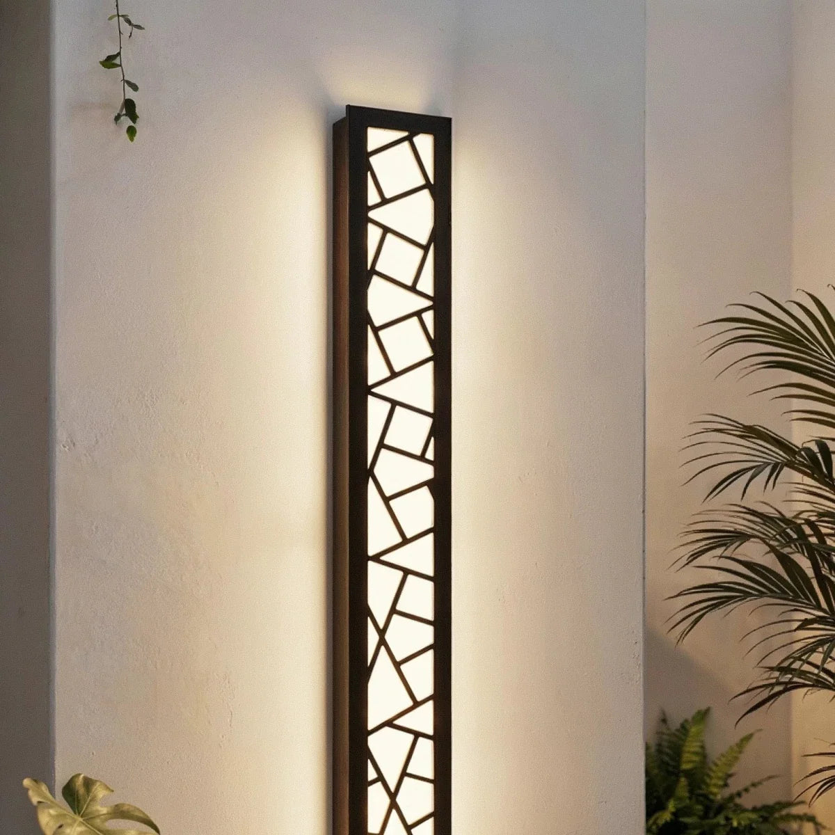 Clérachic tall black wall lamp with a geometric cut-out grille glowing warm white on a white wall among palm leaves.