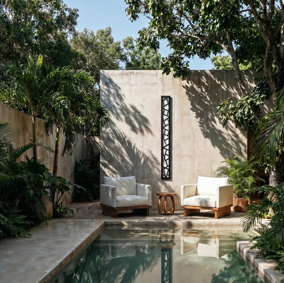Clérachic tall black cut-out wall lamp on a concrete garden wall above two cream armchairs, beside a plunge pool.
