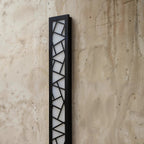 Clérachic close view of a black aluminium wall lamp, its geometric cut-out grille over a white diffuser on weathered concrete.