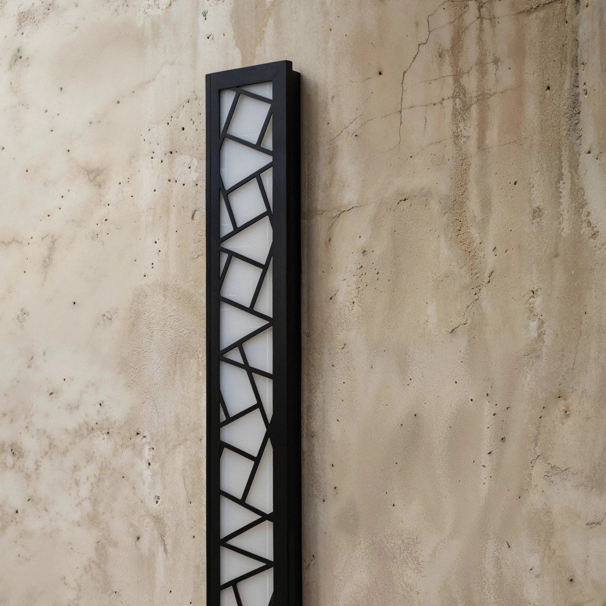Clérachic close view of a black aluminium wall lamp, its geometric cut-out grille over a white diffuser on weathered concrete.