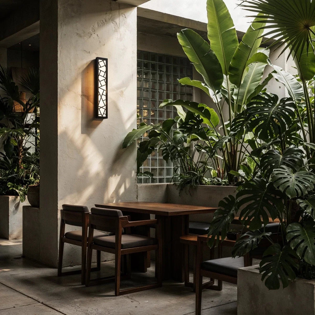 Clérachic black cut-out wall lamp glowing amber on a pale terrace wall above a wooden table and chairs among tropical plants.