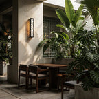 Clérachic black cut-out wall lamp glowing amber on a pale terrace wall above a wooden table and chairs among tropical plants.