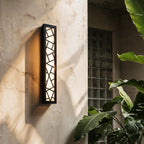 Clérachic black aluminium wall lamp with a geometric cut-out grille, lit warm and casting a shadow on a rough plaster outdoor wall.