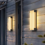Two Clérachic ribbed wall lights, one small and one tall, glowing warm on a grey clapboard wall beside a doorway.
