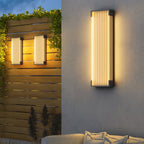 Tall Clérachic wall light on a rendered wall, with two smaller ones lit on a slatted timber fence behind.