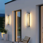 Pair of tall ribbed wall lights from Clérachic on a white rendered house wall at dusk, beside a glass door.