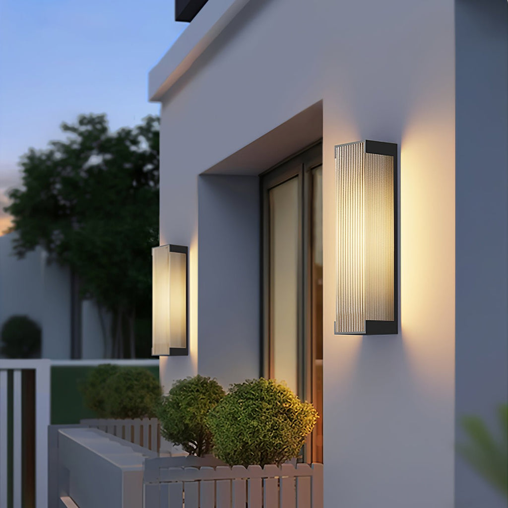 Lit Clérachic wall lights on a white house front at dusk, with clipped shrubs and trees in the garden below.