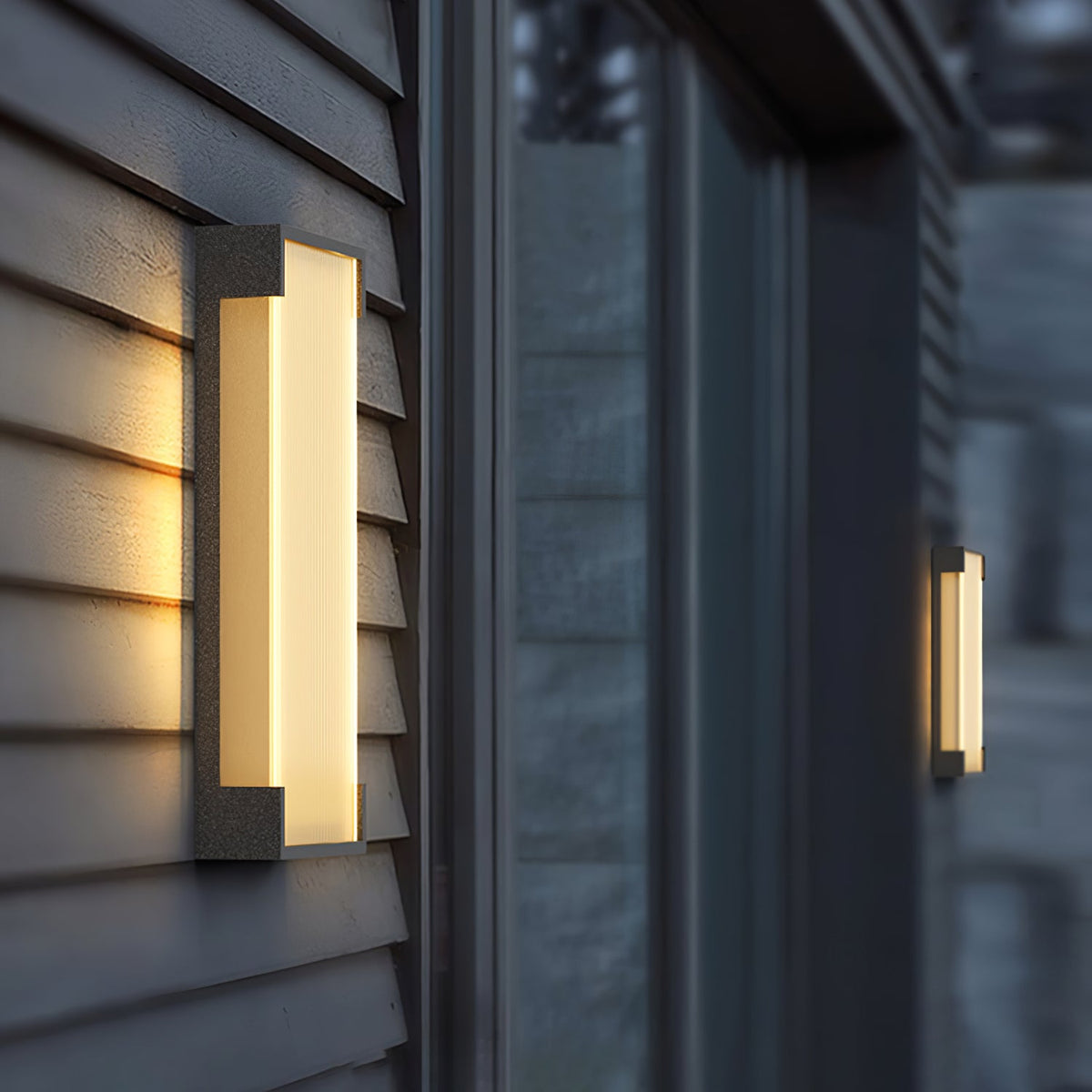 Soft-focus view of a deep Clérachic wall light on grey cladding, with a second light blurred beyond a window.