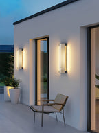 Two ribbed wall lights from Clérachic either side of a tall window on a white wall, with a chair and potted plants.