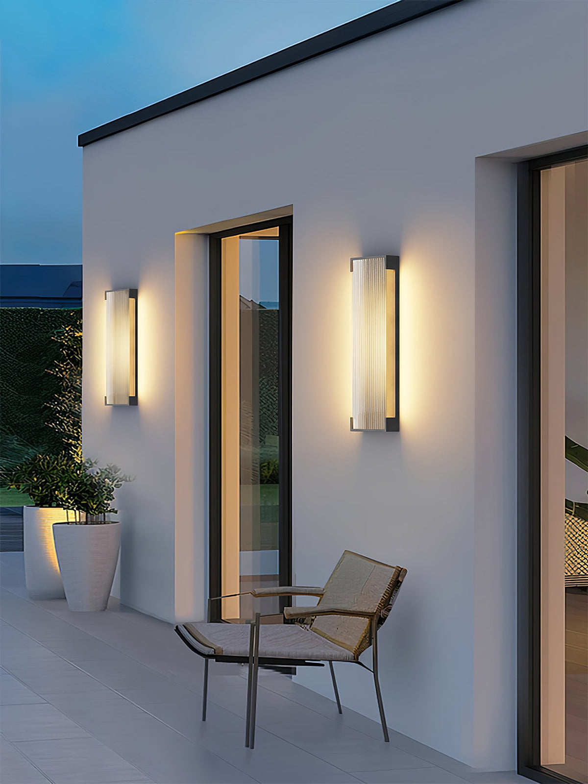 Two ribbed wall lights from Clérachic either side of a tall window on a white wall, with a chair and potted plants.
