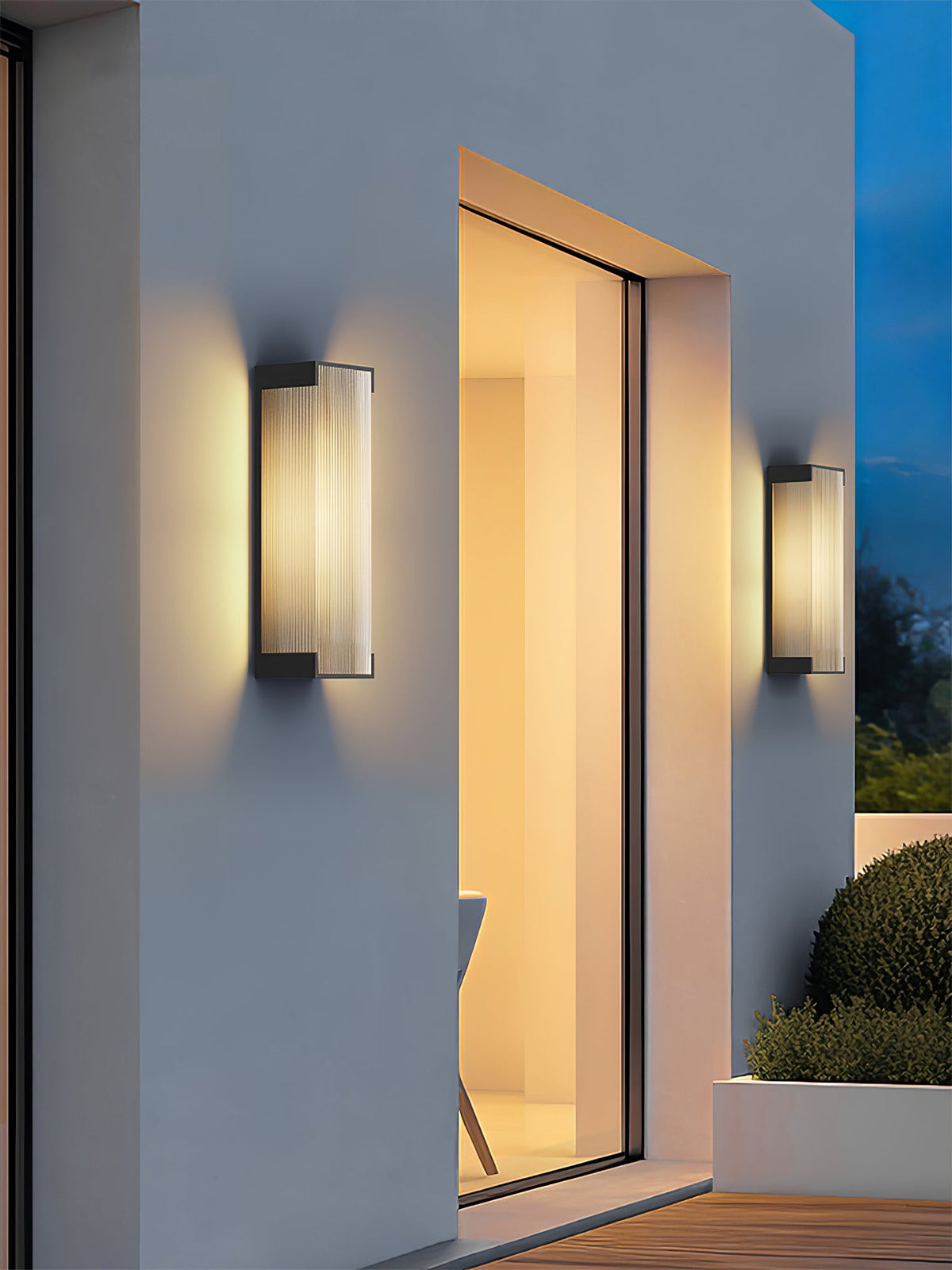 Pair of Clérachic wall lights framing a recessed doorway on a white wall, with shrubs and evening sky around.