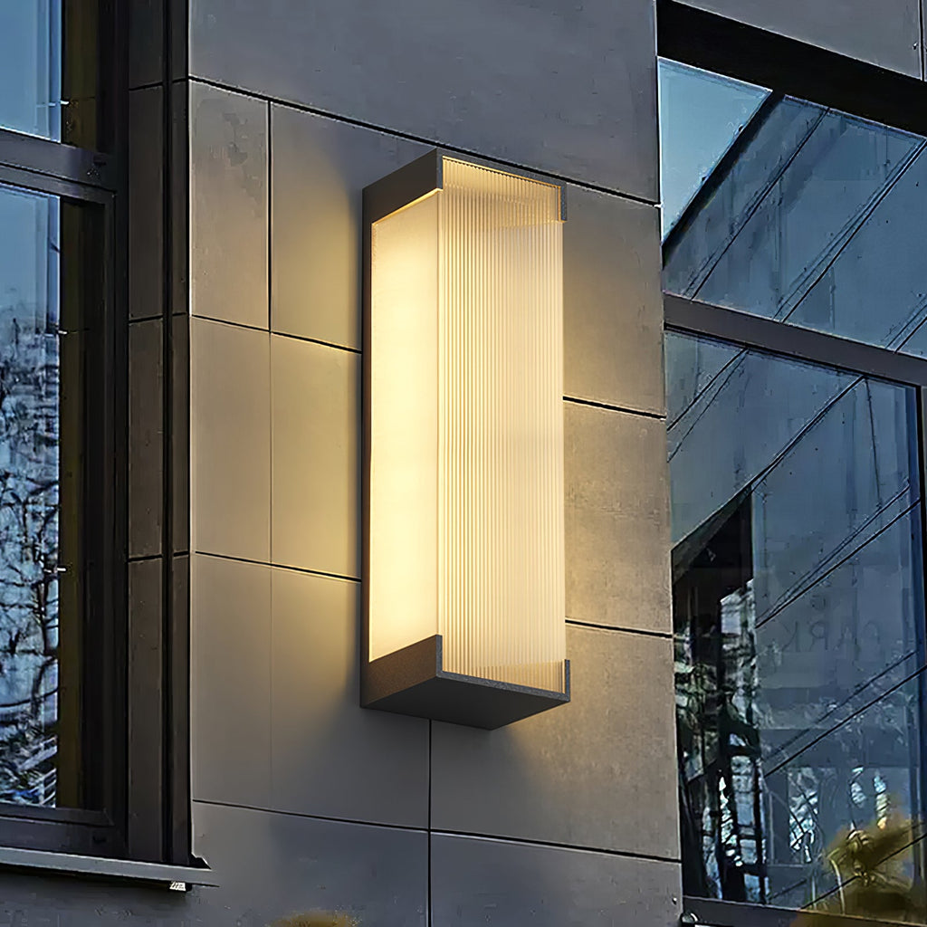 Close view of a Clérachic rectangular wall light, ribbed panel lit warm, on a grey tiled wall beside a window.