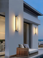 A pair of Clérachic wall lights on a white house corner, above a wicker sofa with grey cushions and a pool edge.