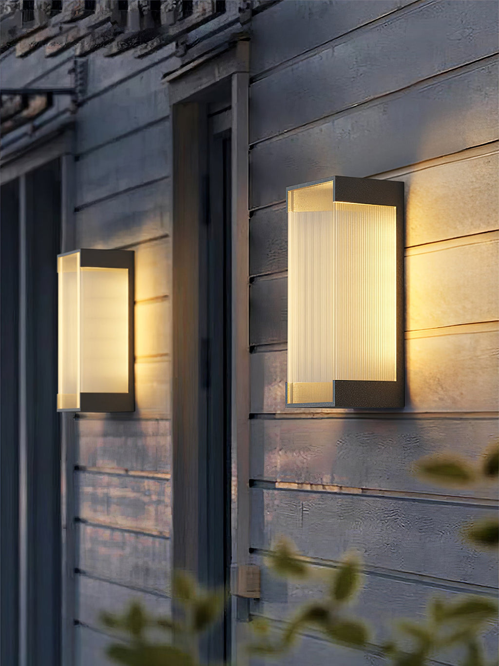 Two Clérachic wall lights with deeper bodies on grey weatherboard cladding, lit warm beside a dark door frame.