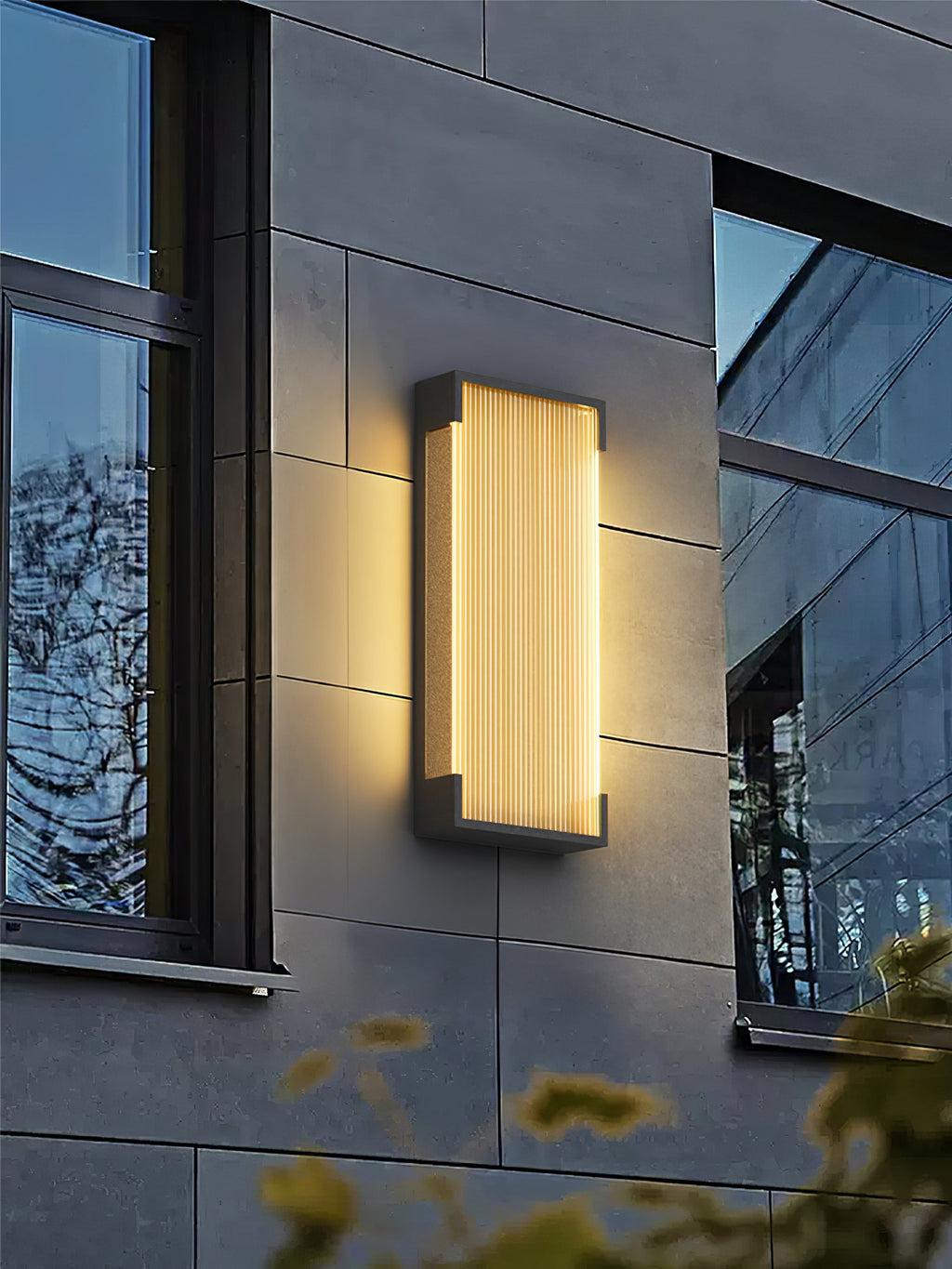 A single Clérachic wall light glowing on a grey panelled facade, between two dark-framed windows.