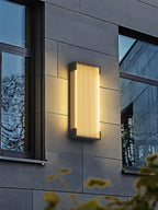 A single Clérachic wall light glowing on a grey panelled facade, between two dark-framed windows.