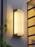 Clérachic wall light seen from slightly below, its ribbed panel lit warm, with a leaf-covered window frame beside it.