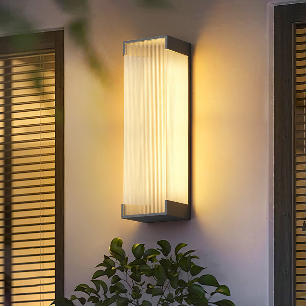 Warmly lit Clérachic wall light on a cream wall beside a slatted shutter, with green leaves in the foreground.