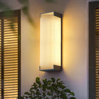 Warmly lit Clérachic wall light on a cream wall beside a slatted shutter, with green leaves in the foreground.
