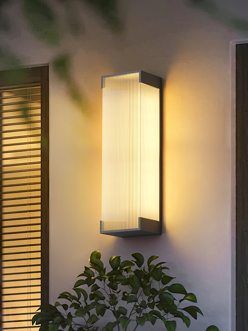 Clérachic ribbed wall light glowing on a cream wall next to wooden shutters, with a leafy shrub below it.