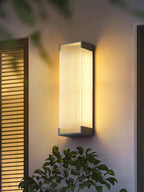 Clérachic ribbed wall light glowing on a cream wall next to wooden shutters, with a leafy shrub below it.