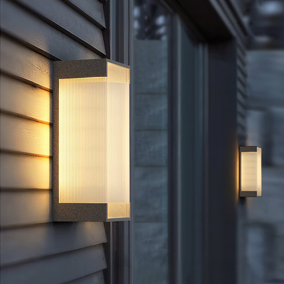 Close view of a deeper Clérachic wall light on grey clapboard, its ribbed panel lit warm, with a second light beyond.