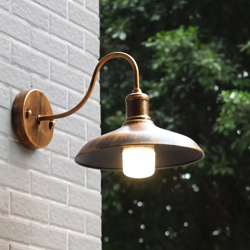 Clérachic bronze gooseneck wall lamp lit on a white brick wall, with garden foliage behind it