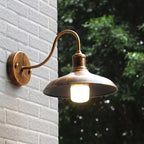 Clérachic bronze gooseneck wall lamp lit on a white brick wall, with garden foliage behind it