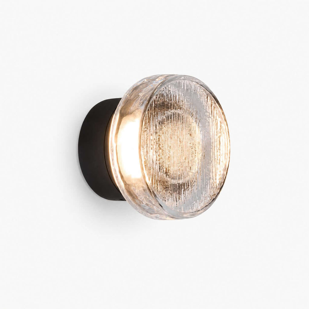 Round outdoor wall light with a clear ribbed glass diffuser on a black backplate, lit warm, on a plain white backdrop, from Clérachic.