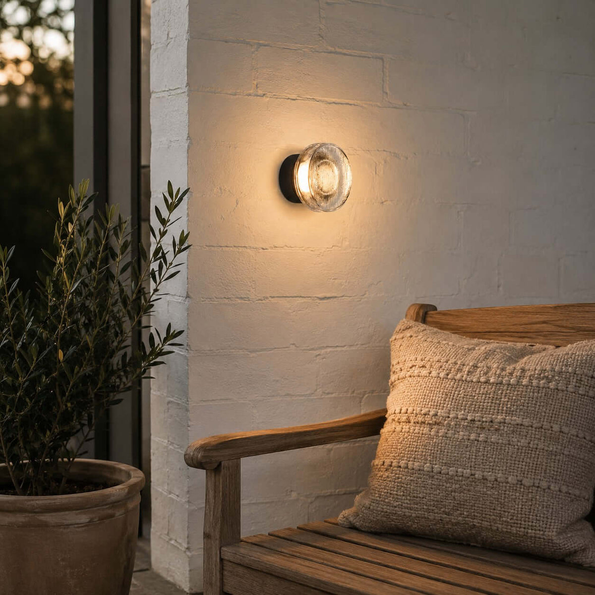 Small round Clérachic outdoor wall light on a white-painted brick wall at dusk, beside a wooden bench and a potted olive.