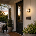 Round Clérachic outdoor wall light beside a black glazed front door, with a planter of hydrangeas and a porch bench.