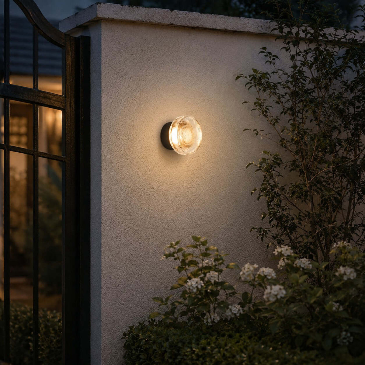 Clérachic outdoor wall light on a rendered garden wall, with white flowering shrubs below and a dark-framed window beside it.