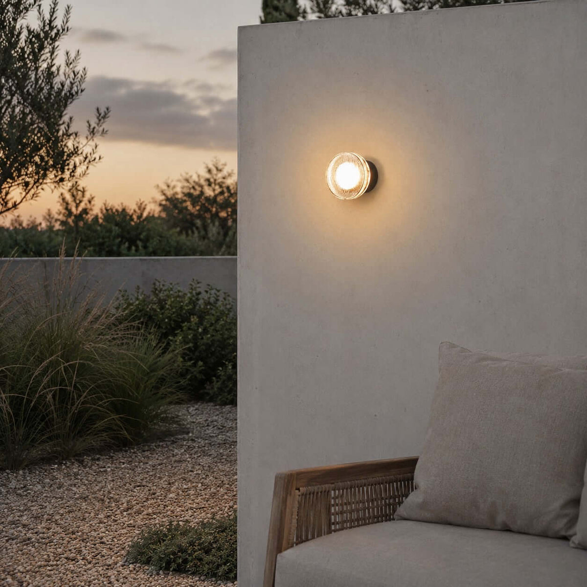 Shown here, the Clérachic outdoor wall light on a plain terrace wall at dusk, with a gravel garden and a rattan sofa at the side.