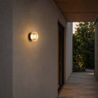 In this photo, the Clérachic outdoor wall light on the wall of a covered walkway, with a timber soffit and a lit garden path beyond.