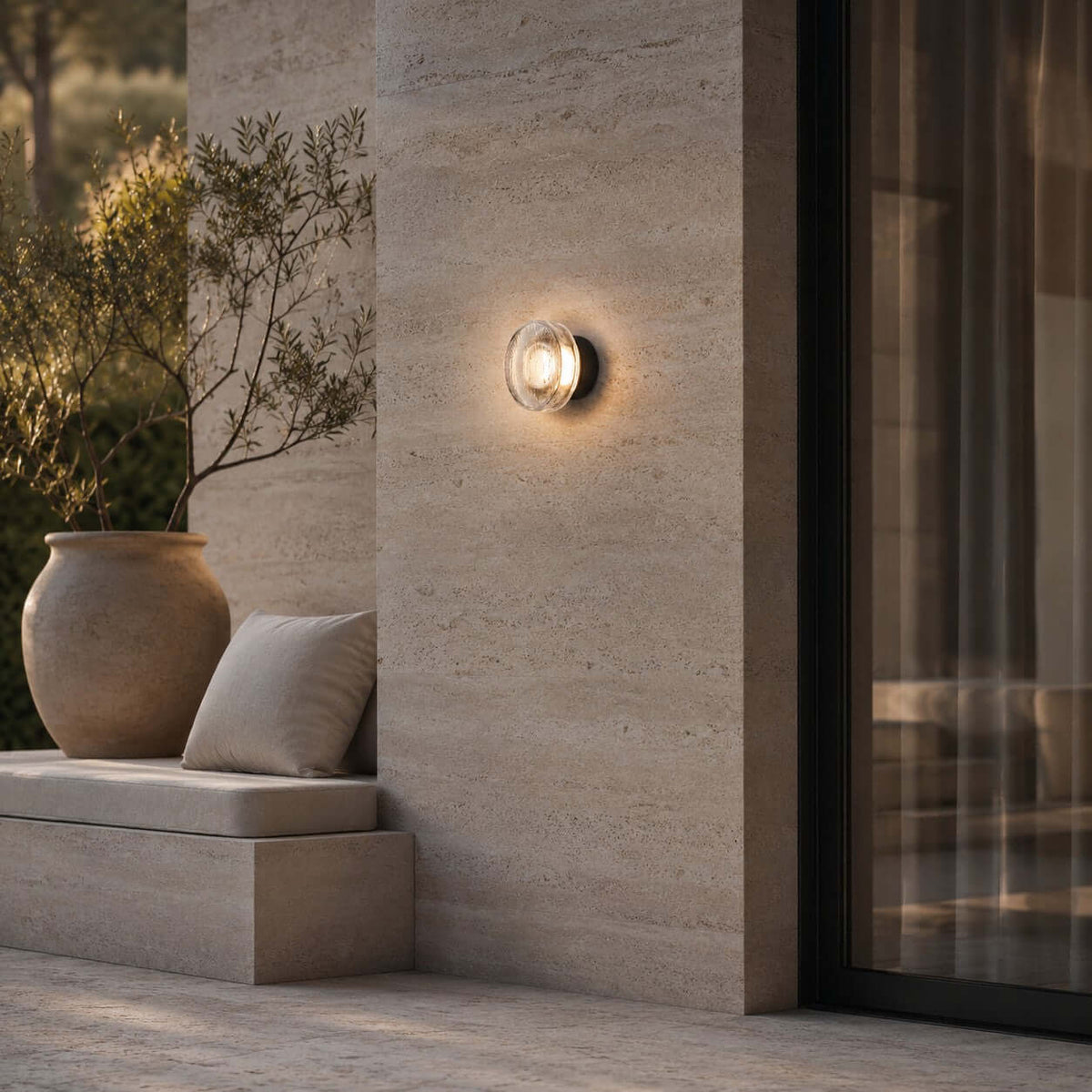In this photo, the Clérachic outdoor wall light on a travertine-clad pier, with a stone bench, a cushion and a large pot with an olive branch.