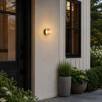 Clérachic outdoor wall light between a black front door and a dark-framed window, with potted herbs and hydrangeas below.