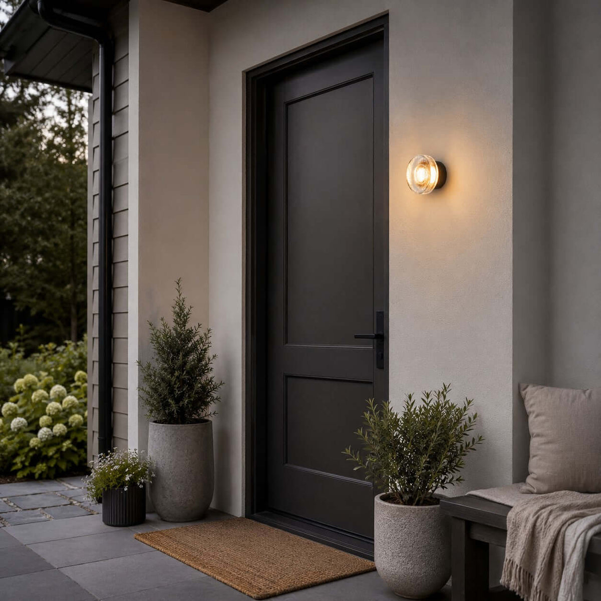 Clérachic outdoor wall light beside a charcoal front door, above a coir doormat and potted evergreens in grey planters.