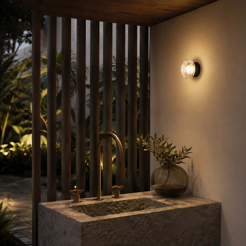 Clérachic outdoor wall light above a stone basin with a brass tap, beside a timber slat screen in a lit garden.
