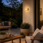 Pictured here, the Clérachic outdoor wall light on a terrace pillar, with a wooden sofa, coffee table and potted plants in the evening garden.