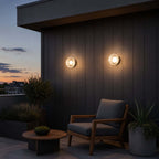 Two Clérachic outdoor wall lights on dark vertical cladding, above a wooden armchair and a round side table at sunset.