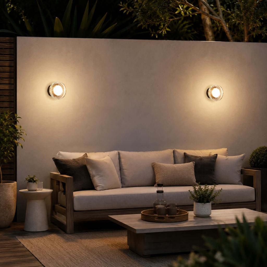 Two Clérachic outdoor wall lights above a garden sofa with scatter cushions and a low coffee table on a timber deck.