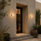 Two Clérachic outdoor wall lights flanking a black-framed glass door in a rendered wall, with stone steps and olive trees.